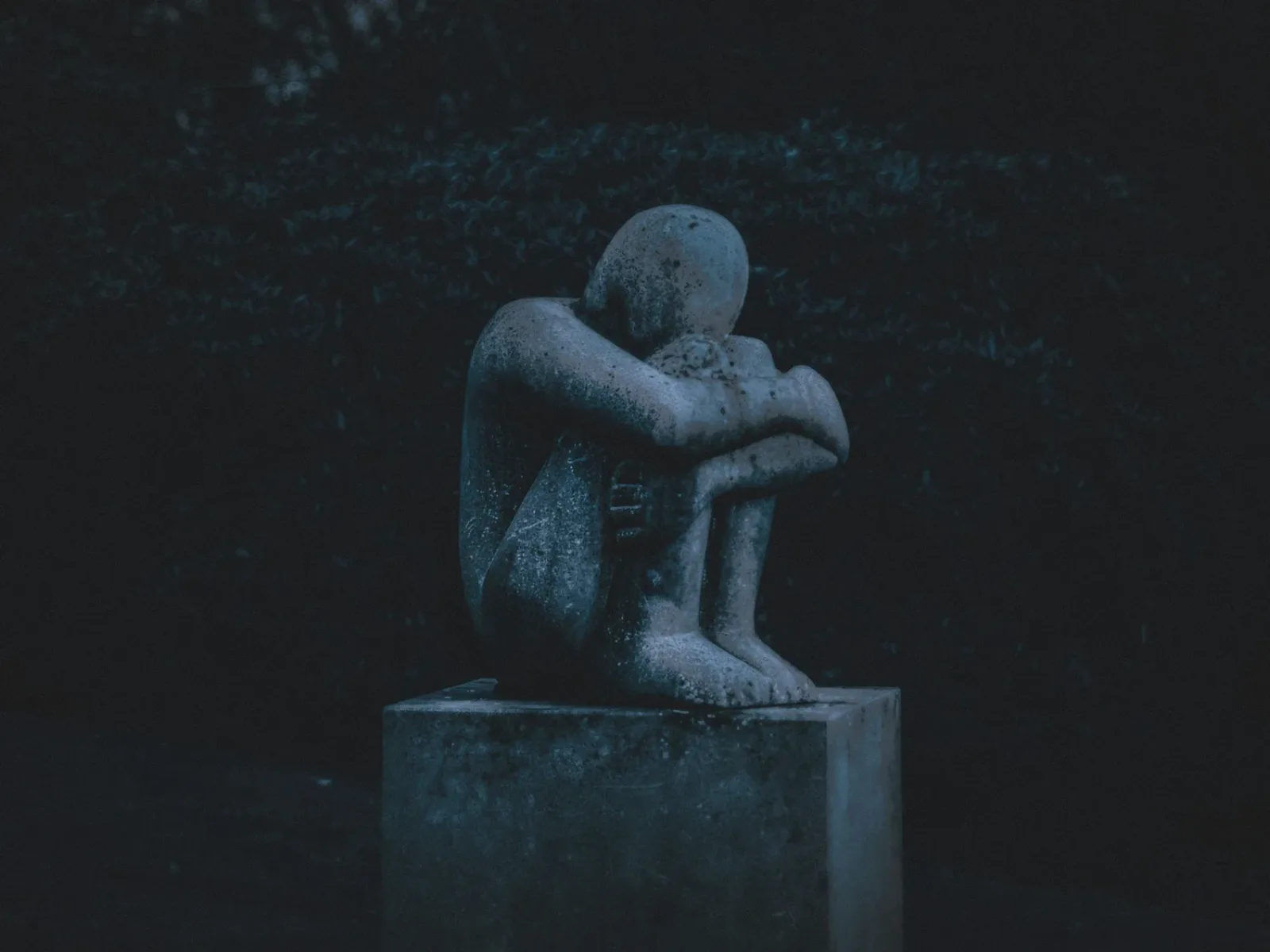 Skulptur av en människa som sitter hopkurad med armarna om knäna, i mörkt skymningsljus